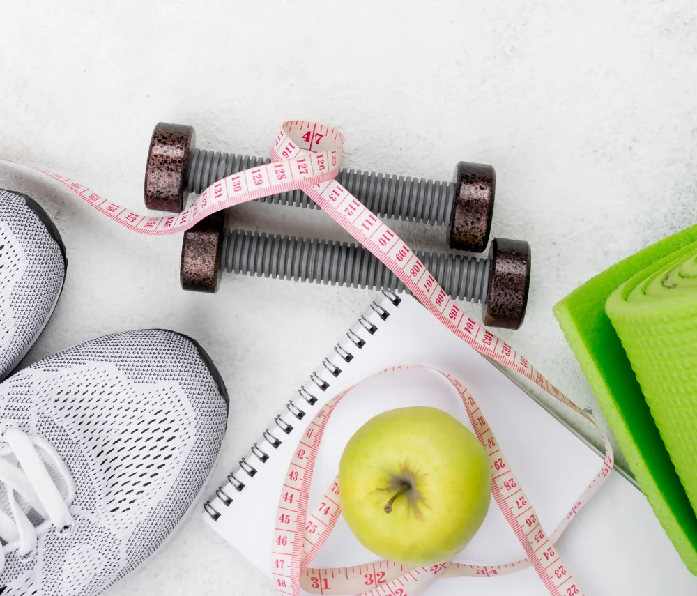 La imagen muestra tenis, pesas, una cinta métrica, una manzana y un tapete de yoga, relacionados con la medicina del deporte. La imagen muestra tenis, pesas, una cinta métrica, una manzana y un tapete de yoga, relacionados con la medicina del deporte.