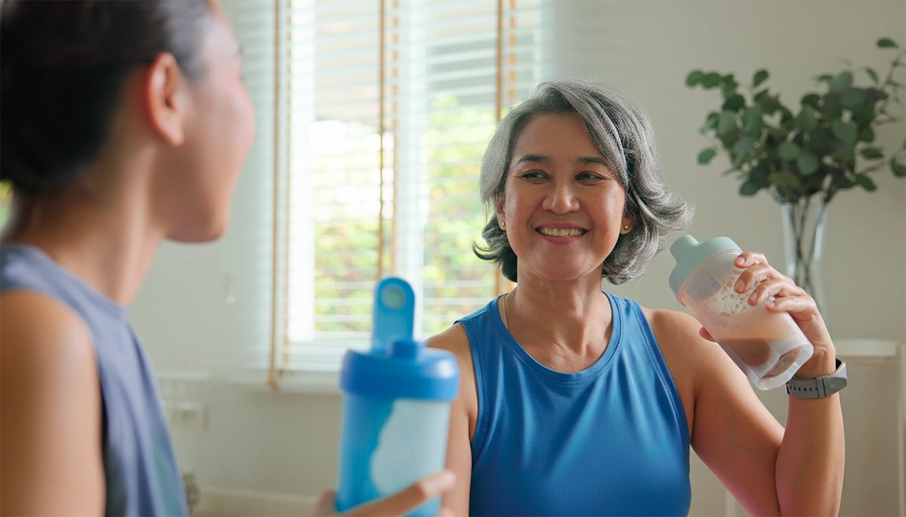 Mujer de mediana edad tomando un batido de proteína, mientras le sonríe a su hija.